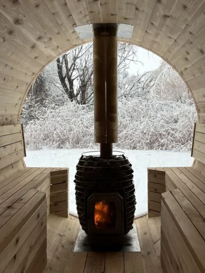 Glowing wood-burning stove inside a barrel sauna with snowy trees visible through a panoramic arched window