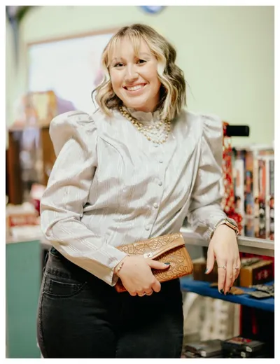Smiling woman in a silver button-up blouse holding a tooled leather clutch in a vintage shop