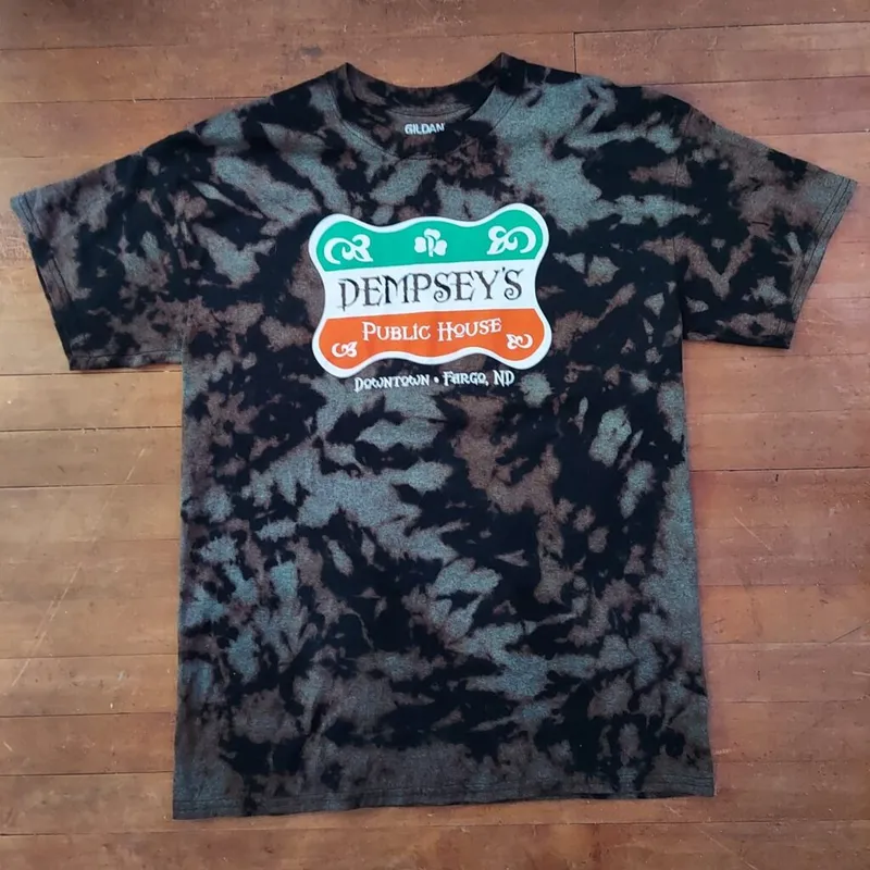 Black acid-washed tie-dye T-shirt with Dempsey's Public House Downtown Fargo ND graphic, laid flat on wood floor