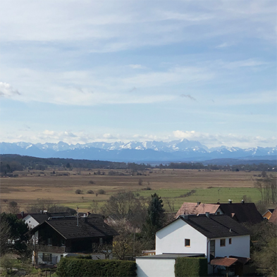 Rural village with dark-roofed houses overlooking flat farmland and snow-capped mountains beyond