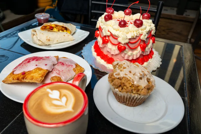 Latte, glazed pastries, crumb muffin, cherry-topped cake, and a breakfast burrito on a cafe table