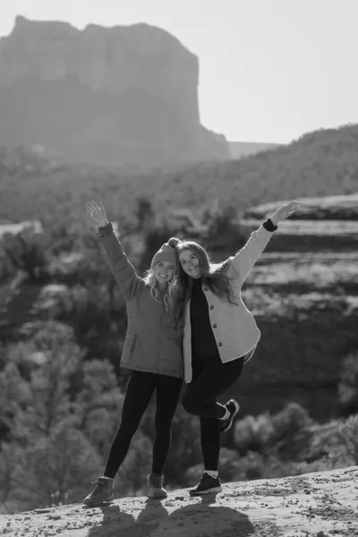 Two smiling women in winter jackets cheering with arms raised on a rocky overlook with mesa backdrop
