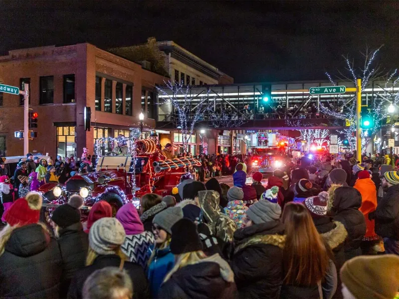 Crowd in winter gear lines a downtown street during a nighttime holiday parade with a lit vintage fire truck