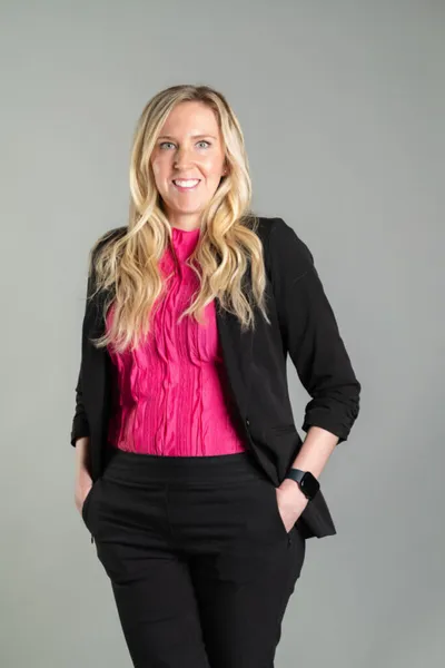 Smiling blond woman in a pink blouse and black blazer, hands in pockets, against a gray backdrop