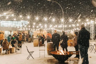 Crowd gathered around a fire pit at an outdoor winter event under string lights in snowfall