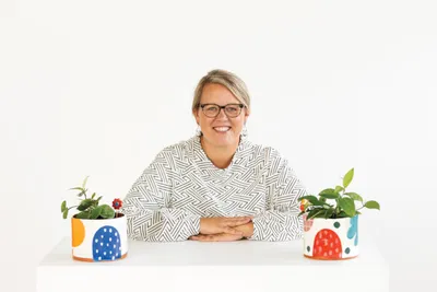 Smiling woman with glasses seated at a white table flanked by two colorful ceramic planters with small plants