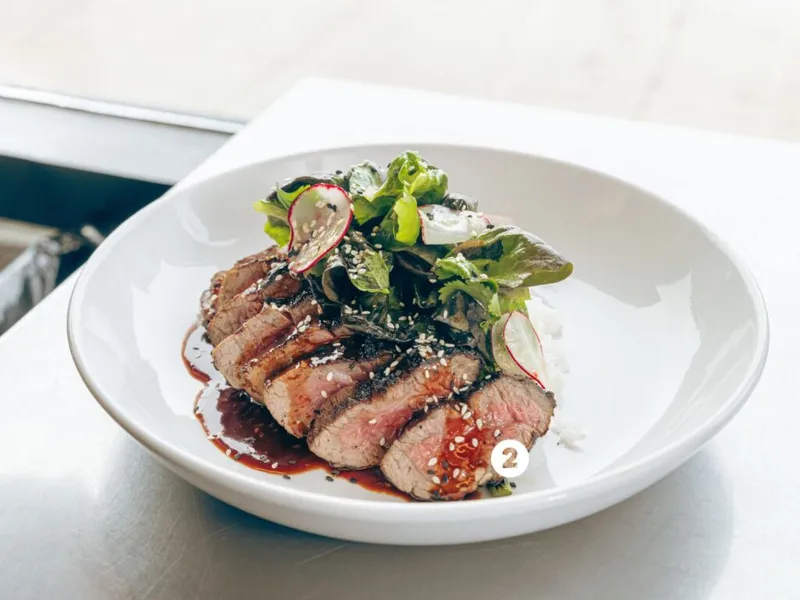 Sliced medium-rare steak over rice with greens, radish, sesame seeds, and dark sauce in a white bowl