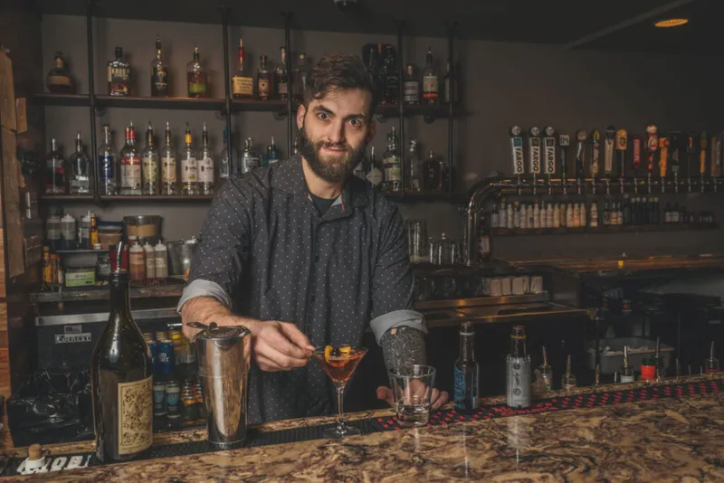 Bearded bartender garnishing a cocktail in a coupe glass behind a well-stocked bar