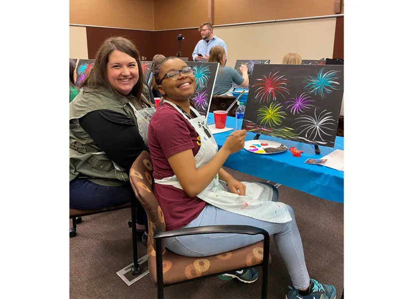 Two smiling women in aprons pose at a group painting class with colorful fireworks canvases