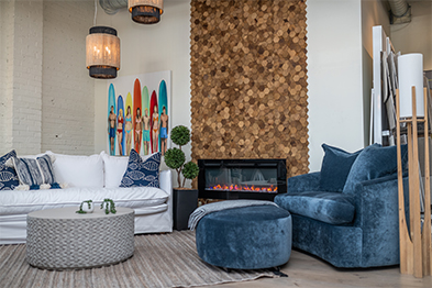 Coastal-styled living room with white sofa, blue velvet chair, wood-tile fireplace, and surfboard artwork