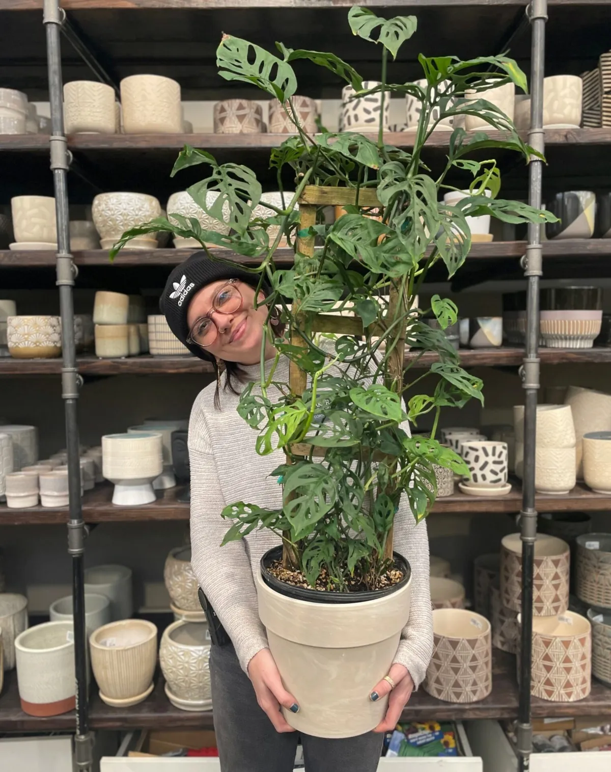 Woman in black beanie holding a large monstera plant in front of shelves of ceramic pots