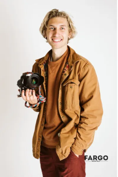 Smiling young man with blonde hair holding a camera, wearing a tan jacket