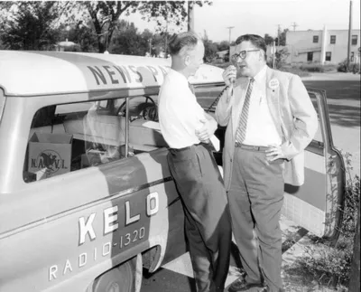 Two men talking beside a KELO Radio 1320 news van in a black-and-white photo