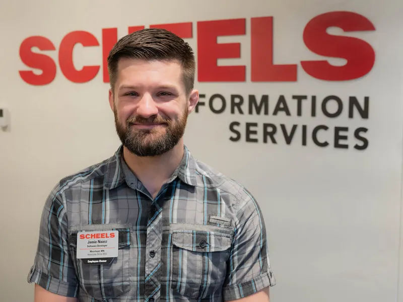 Smiling bearded man wearing a Scheels name badge in front of a Scheels Information Services sign