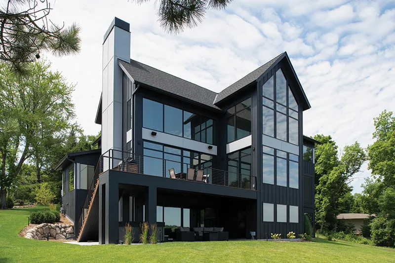 Dark modern two-story home with floor-to-ceiling windows, upper deck, and tall chimney surrounded by green lawn and trees