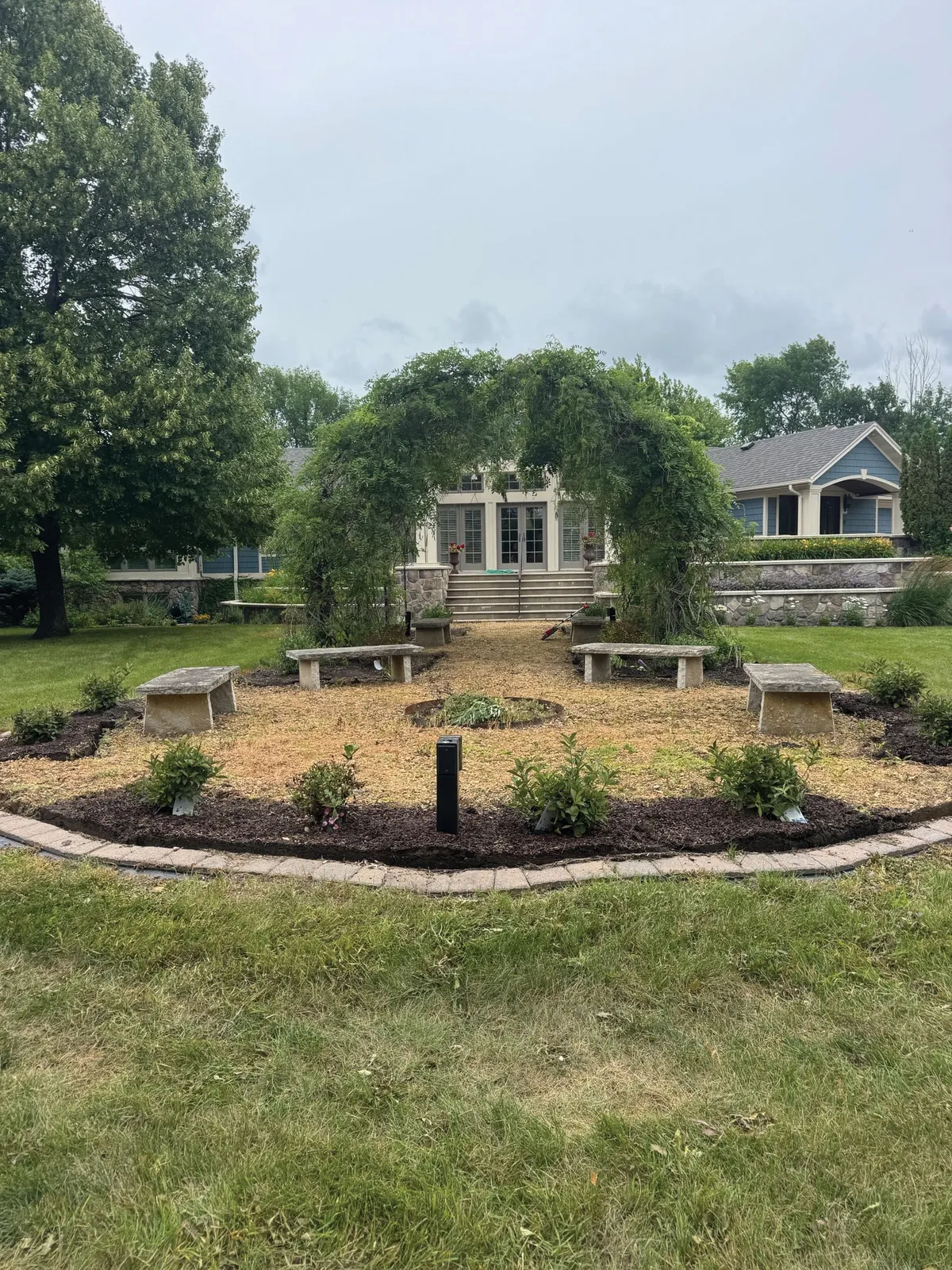 Circular mulched garden with stone benches and a vine-covered arbor leading to a house entrance