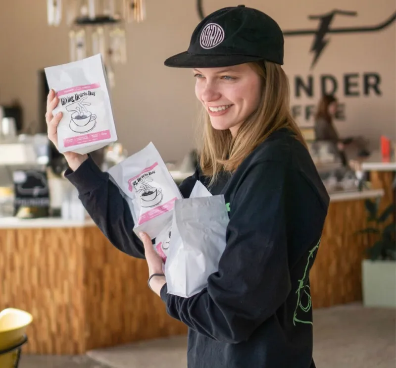 Smiling woman in black cap holding white Giving Hearts Day coffee bags inside a café