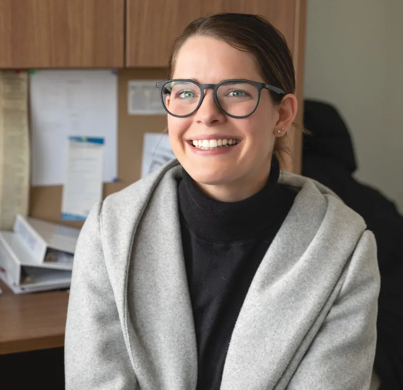 Smiling woman with glasses and gray wrap seated at an office desk