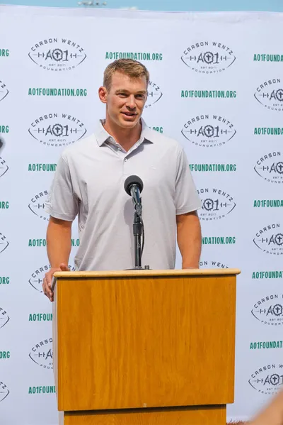 Man in gray polo speaks at a wood podium before an AO1 Foundation branded backdrop