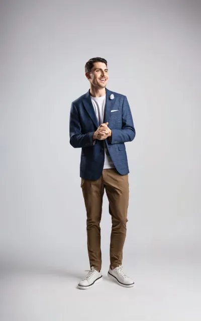 Smiling man in navy blazer, white tee, tan chinos, and white sneakers on gray background