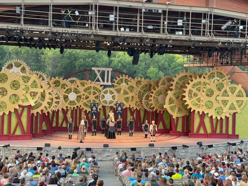 Costumed performers on an outdoor stage with giant golden gear set pieces before a large audience