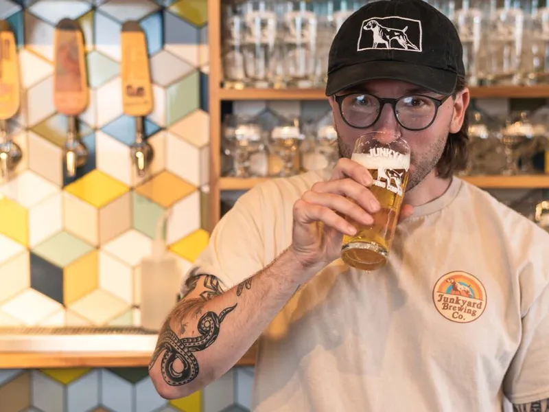 Tattooed man in a Junkyard Brewing Co. shirt sipping a pint of beer at a tap room bar