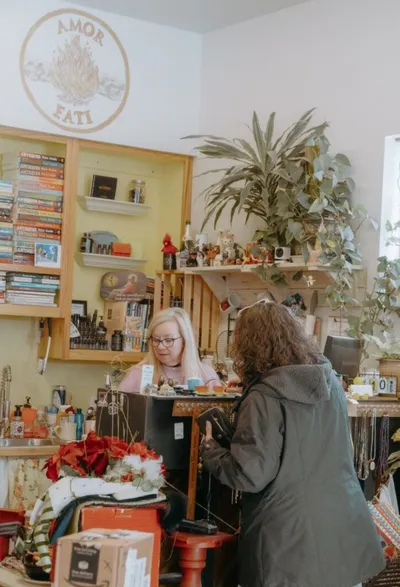 Two women at a cluttered shop counter beneath an 'Amor Fati' wall sign, surrounded by books and plants