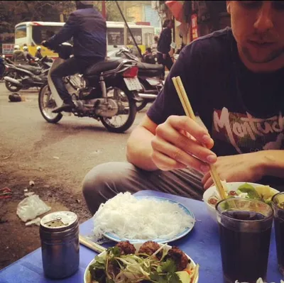 Man eating noodles with chopsticks at a street-side table, motorbikes passing behind him