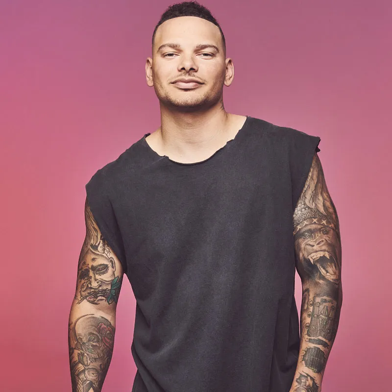 Heavily tattooed man in a black sleeveless shirt against a pink background