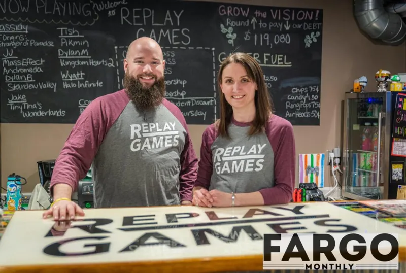 Two smiling staff members in Replay Games shirts stand behind a counter with a large store sign