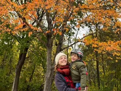 Woman holding a young child in a bike helmet beneath orange autumn trees