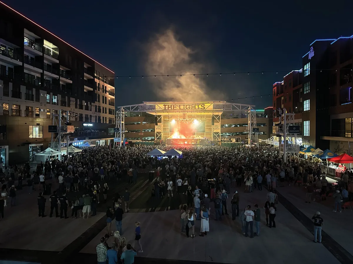 Large crowd fills an outdoor plaza at night facing a lit concert stage at The Lights venue
