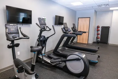 Small fitness room with stationary bike, elliptical, treadmill, wall-mounted TVs, and medicine balls