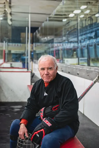 Elderly man in hockey gear sits rinkside holding a helmet and stick