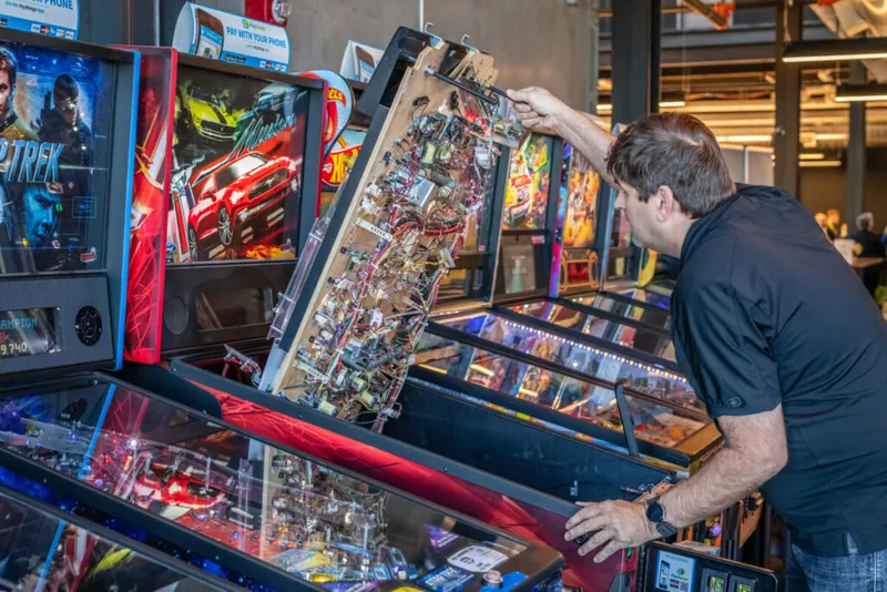 Man lifting the playfield of a pinball machine in a room full of arcade cabinets