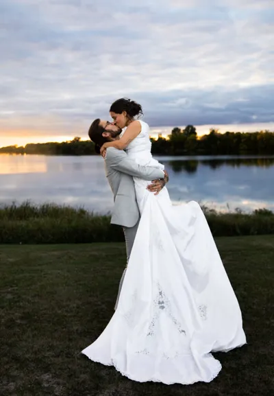 Groom in gray suit lifts bride in white gown at lakeside sunset