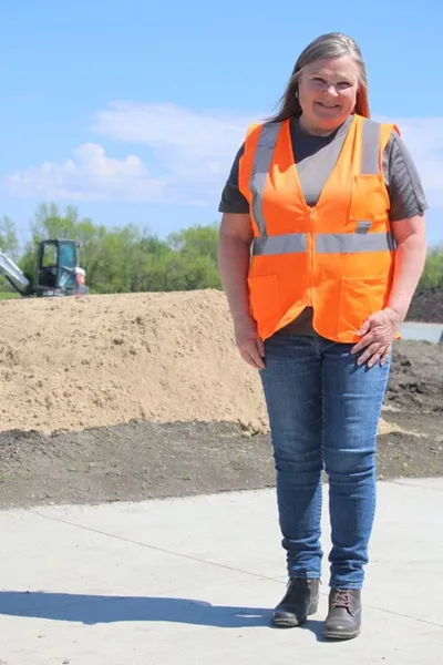 Woman in orange safety vest and jeans stands at an outdoor construction site