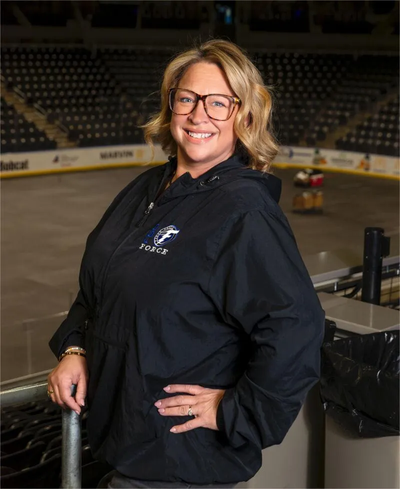 Smiling woman in a black Force jacket leaning on a rail inside an empty hockey arena