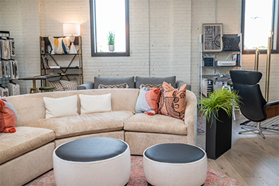 Curved beige sectional sofa with colorful pillows and two round ottomans in a bright showroom