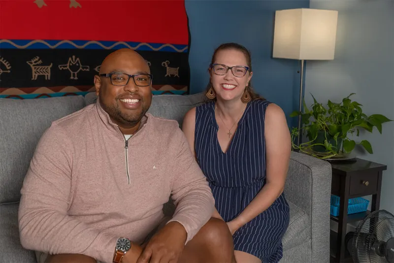 Man in glasses and pink pullover sits smiling beside a woman in a navy dress on a gray sofa