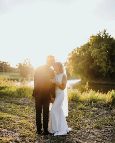 Couple in wedding attire embracing at golden hour beside a calm river