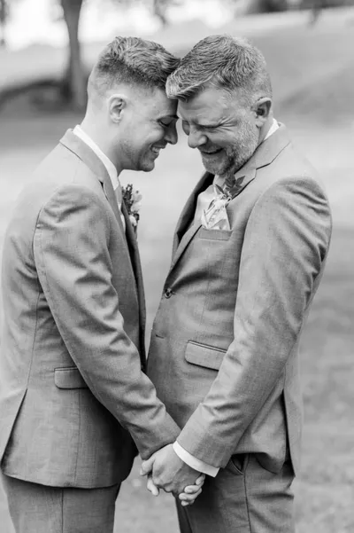 Two grooms in matching gray suits hold hands and touch foreheads, smiling