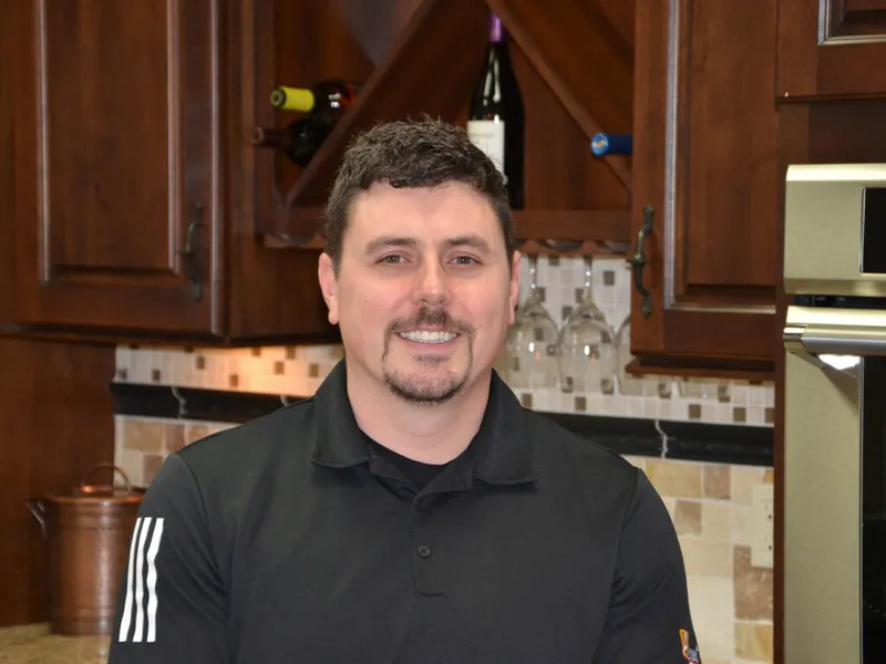 Smiling man with goatee in black polo standing in a kitchen with wine rack cabinetry