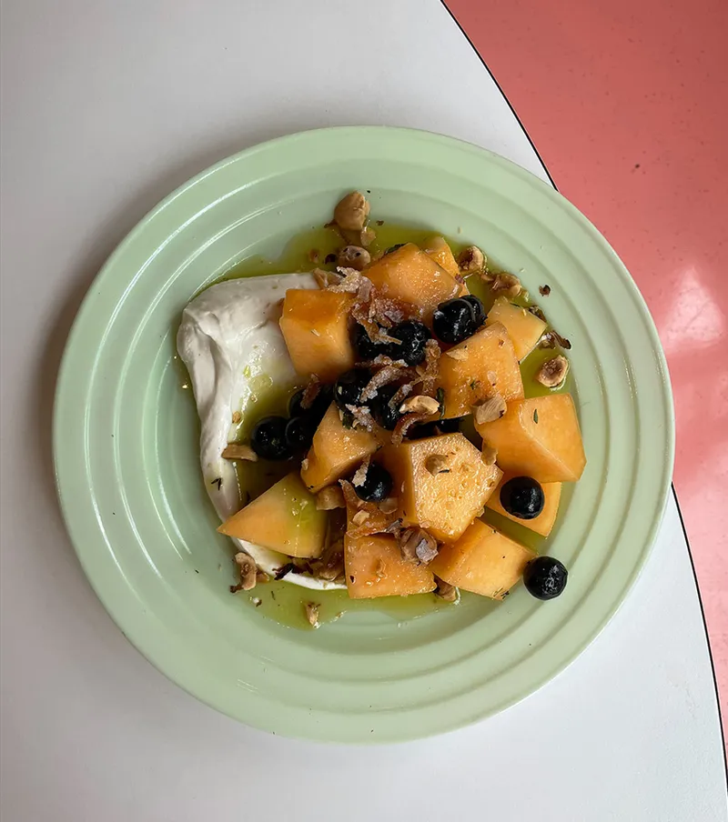 Cubed cantaloupe with blueberries, crushed hazelnuts, and whipped cream on a sage green plate