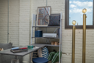 Styled corner with a leaning shelf, desk, patterned pillows, and brass floor lamp against a white brick wall