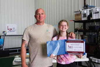 Man and young girl smiling indoors, holding a certificate of completion and a blue folder