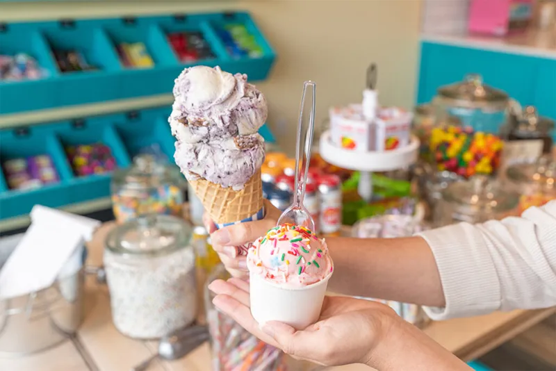 Hands holding a triple-scoop purple ice cream cone and a pink sprinkle cup inside a candy shop