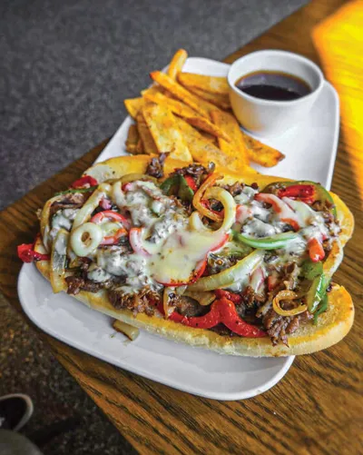 Open-faced cheesesteak sandwich topped with peppers, onions, and melted cheese, served with fries and dipping sauce