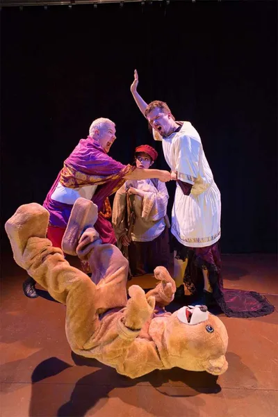 Three costumed stage actors react dramatically over a performer in a bear suit lying on the floor