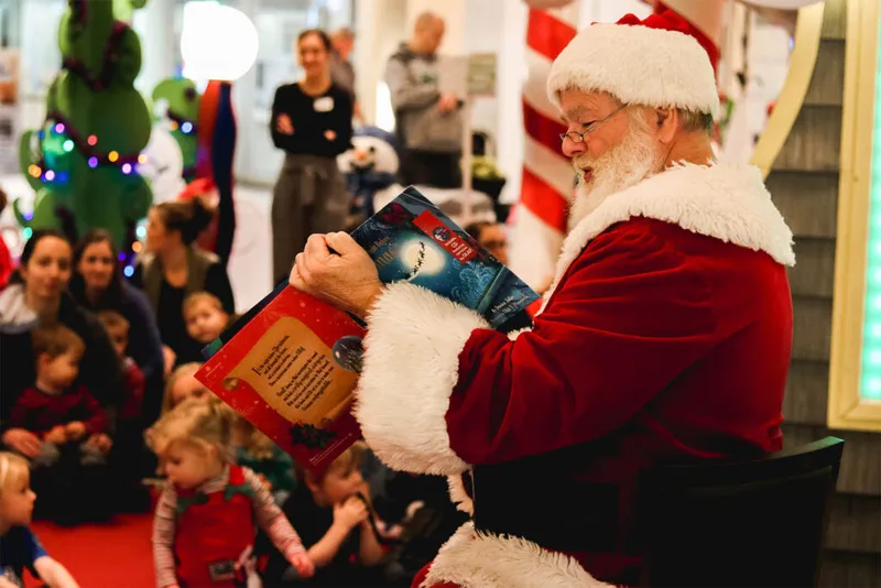 Santa Claus reading a picture book aloud to a crowd of young children indoors